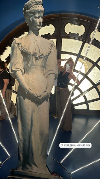 Una mujer rubia se toma una selfie con un teléfono frente a una figura de tamaño real de una mujer con vestido de época, dentro de un museo con ventanales