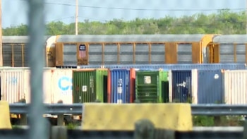 Un vagón de tren de carga Union Pacific, amarillo y gris, se observa sobre una fila de contenedores de envío multicolores. Follaje verde en el fondo