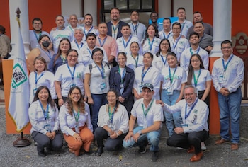 Un grupo de personas posa sonriendo en varias filas frente a un edificio con columnas y una bandera a la izquierda