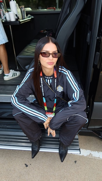 Mujer con cabello largo y oscuro, gafas de sol, chándal negro y azul con franjas blancas, sentada en el estribo de una camioneta. Se observa el pie de otra persona