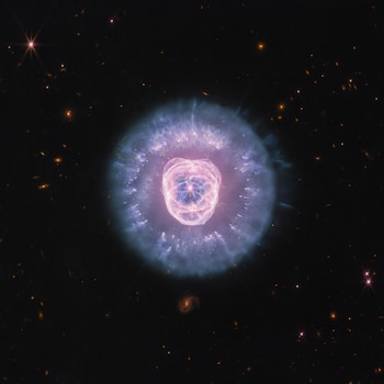 Nebulosa planetaria con un núcleo rosa y azul, anillos de gas y polvo, estrellas y galaxias en el fondo oscuro del espacio