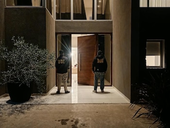 Dos personas con camperas oscuras que llevan la sigla DDI en la espalda están de pie frente a la puerta principal iluminada de una casa