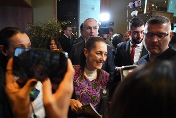 Claudia Sheinbaum sonríe, rodeada de personas y teléfonos móviles. Viste una prenda púrpura y blanca, en un ambiente interior iluminado