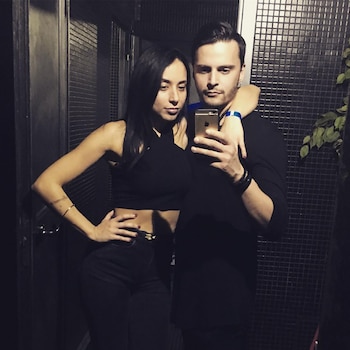 Un hombre y una mujer, ambos con ropa oscura, se toman una selfie con un teléfono dorado en un interior con paredes oscuras y vegetación a la derecha