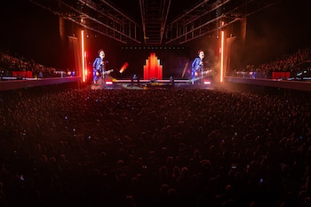 Gran multitud en un concierto con un escenario iluminado en tonos rojos y naranjas. Pantallas gigantes muestran a un guitarrista