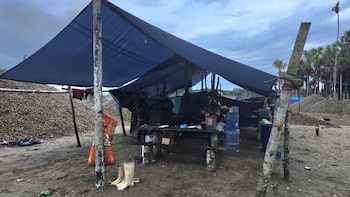 Acampamento rural com telhado azul, postes, mesa, balde de água e sapatos no chão, cercado por seixos e árvores, sob céu nublado
