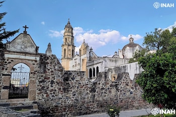 Muro de piedra con una puerta de hierro en arco, una torre de campanario al fondo y una cúpula junto a árboles y cielo despejado