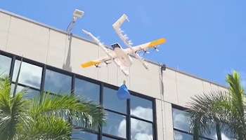 Dron blanco y amarillo con un paquete azul suspendido, volando frente a un edificio beige con grandes ventanas y frondas de palmeras verdes
