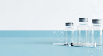 Tres frascos transparentes de medicamento con tapa gris y una jeringa apoyada en una superficie azul claro, con un fondo blanco y patrones hexagonales.