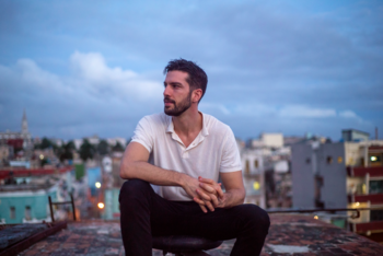 Hombre joven barbudo con camiseta blanca, sentado en una azotea oscura. Edificios borrosos y un cielo nublado al atardecer en el fondo