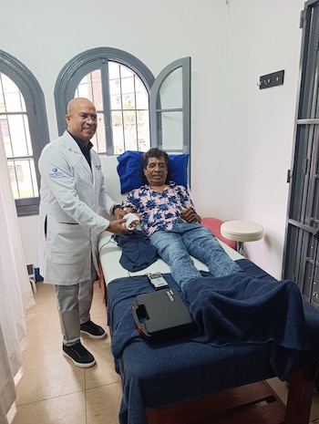 Un doctor de bata blanca revisa la mano vendada de Cachay, quien reposa en una camilla médica. Hay un dispositivo médico y toallas azules sobre la camilla