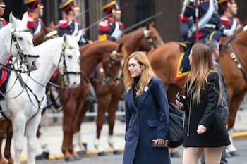 Dos mujeres caminan, una de rubio con abrigo azul marino y la otra con abrigo negro, mientras jinetes uniformados montan caballos blancos y marrones de fondo