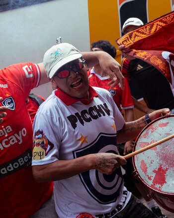 Un hombre con gorra blanca, gafas de sol y camiseta de un equipo de fútbol toca un bombo rodeado por otros aficionados vestidos de rojo