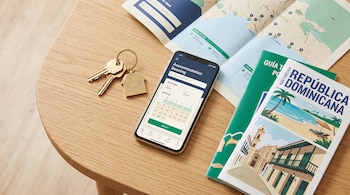 Llaves de apartamento con llavero de casa, un smartphone con una app de reservas y una guía turística de República Dominicana sobre una mesa de madera clara.