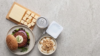 Una hamburguesa en un plato, un tazón de yogur con cereales, trozos de queso sobre una tabla de madera y una lata de metal sobre piedra