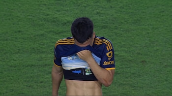 Un jugador de fútbol de espaldas, con camiseta azul y amarilla levantada, muestra una prenda interior clara y un dispositivo de seguimiento en su torso. Fondo de césped verde.
