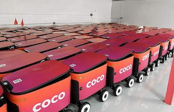 Filas de robots de entrega Coco, de color naranja y magenta, con el logo "COCO" en el costado, alineados en un almacén