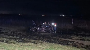 Un automóvil destruido en un campo oscuro junto a tres personas, una de ellas con chaleco reflectante y linterna