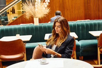 Mujer joven de cabello castaño sentada en un restaurante, mirando su teléfono móvil. En la mesa hay una taza de café y documentos