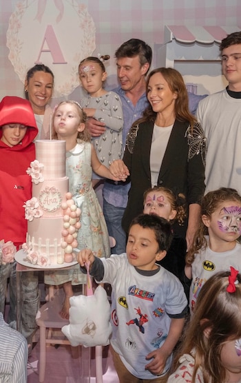Adultos y niños sonríen alrededor de una torta de cumpleaños rosa de tres pisos; algunos niños tienen caras pintadas y uno sostiene algodón de azúcar