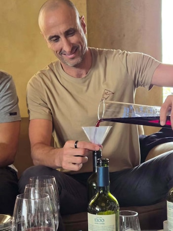Manu Ginóbili vierte líquido rojo desde un recipiente graduado hacia una botella con un embudo, rodeado de copas y botellas