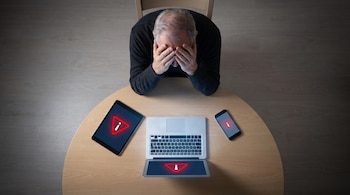 Un adulto mayor con las manos en el rostro está sentado frente a una mesa redonda con una tableta, una computadora portátil y un teléfono móvil que muestran íconos de advertencia.