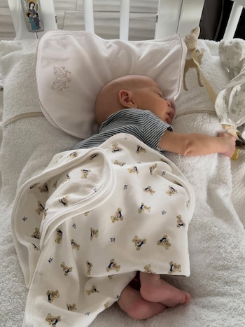 Un bebé duerme de lado en una cuna blanca, envuelto en una manta blanca con estampado de ositos y con un chupete en la boca; sus pequeños pies rosados son visibles