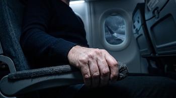 Una mano masculina apretada sobre el apoyabrazos de un asiento de avión, con una ventanilla redonda al fondo que muestra un cielo nublado.