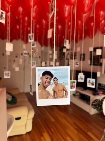 Una habitación llena de globos rojos en forma de corazón y decenas de fotografías pequeñas colgando de cuerdas, con una imagen grande de dos hombres en la playa al frente