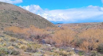 Arroyo que fluye entre vegetación arbustiva seca y laderas de colinas frente a un cielo azul con nubes