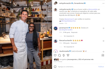 Un hombre sonriente con uniforme de chef y una mujer sonriente con gafas y cabello trenzado posan en una cocina con estanterías y un mostrador de alimentos