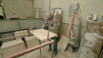Un trabajador con gorra, protectores auditivos y mascarilla corta una losa de piedra artificial sujeta a una mesa de trabajo en un taller polvoriento