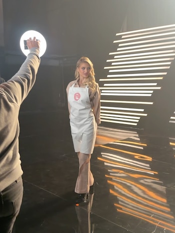 Una mujer rubia con delantal blanco posa en un estudio de televisión frente a un aro de luz sostenido por una persona