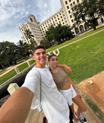 Dos jóvenes sonríen en primer plano, con un gran edificio blanco y verde césped con palmeras al fondo bajo un cielo nublado