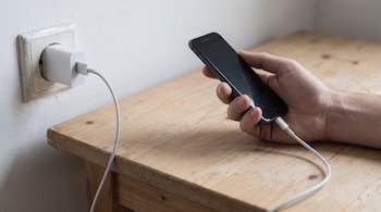 Una mano sostiene un teléfono móvil negro enchufado con un cable blanco a un cargador de pared blanco en un tomacorriente blanco sobre una mesa de madera clara.