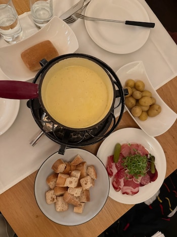 Vista aérea de una mesa de madera con una fondue de queso, pan en cubos, patatas pequeñas, y un plato de embutidos con pepinillos