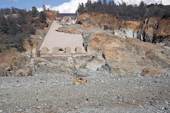 Canal de hormigón fracturado sobre una ladera rocosa erosionada, con una máquina excavadora y cuatro personas con chalecos en la base