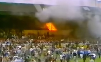 Incendio masivo en las gradas de un estadio, con llamas intensas y denso humo gris. Una gran multitud de personas se aglomera en el césped del campo