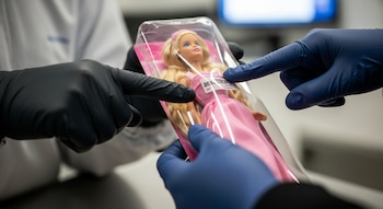 Primer plano de dos pares de manos enguantadas, uno en negro y otro en azul, inspeccionando una muñeca Barbie rubia dentro de su empaque transparente.