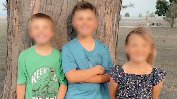 Tres niños de pie frente a un gran tronco de árbol, con sus rostros pixelados. El niño de la izquierda lleva una camiseta verde, el del centro una azul y la niña un top floral.
