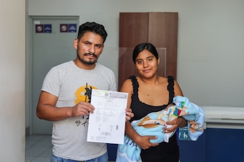 Un hombre, una mujer y un bebé envuelto en una manta azul posan de pie en el interior de una sala; el hombre sostiene un documento en sus manos