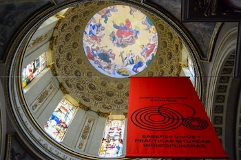 Un gran cartel rojo con texto blanco y círculos negros cuelga bajo una cúpula ricamente decorada con frescos celestiales y ventanas de vitral