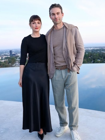 Leighton Meester, con blusa y falda negras, y Chace Crawford, con camisa y chaqueta beige y pantalones claros, posan con una piscina y la ciudad detrás