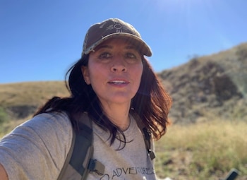 Retrato de una mujer de mediana edad con cabello oscuro y gorra de camuflaje, mirando a la cámara, con un paisaje seco de colinas y cielo azul brillante de fondo