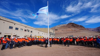 Decenas de personas con cascos y chalecos naranjas observan una bandera celeste y blanca flameando en un mástil en un entorno montañoso