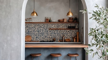 Interior de cocina/bar moderno con arco. Presenta una barra de madera, tres taburetes, azulejos azules y beige, estantes con cristalería y lámparas doradas. Planta verde a la derecha.