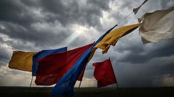 Varias banderas de tela en tonos amarillo, azul y rojo ondean con fuerza bajo un cielo nublado y oscuro. Un rayo de luz blanca desciende de las nubes.