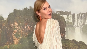 Inés Estevez de espaldas con un vestido blanco de flecos y escote bajo, mirando por encima del hombro. Fondo de montañas rocosas y cascadas