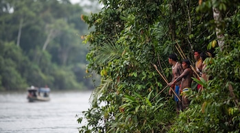 Un grupo de hombres indígenas con pintura corporal y arcos, flechas y lanzas, de pie en la orilla de un río, con densa vegetación tropical y un bote a distancia.