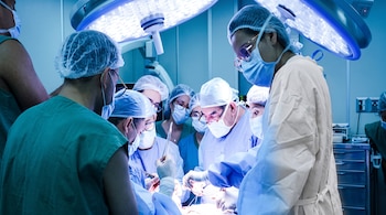 Un equipo de médicos con mascarillas, gorros quirúrgicos y vestimenta azul opera a un paciente bajo luces de quirófano